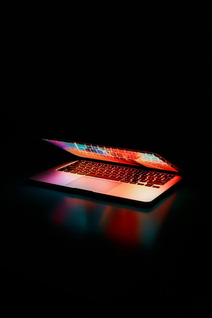 pexels-photo-2047905-2047905 Laptop partially opened, emitting vibrant multicolored lights in dark setting.